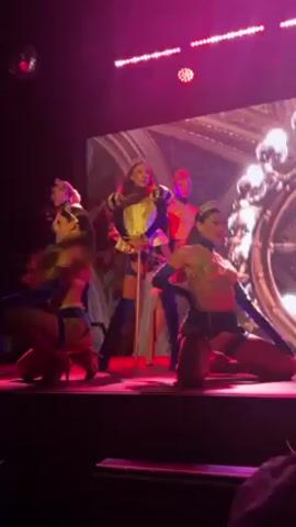 Here's a description of the video:

On a stage lit with red and purple, five dancers perform. The central figure is dressed in a costume reminiscent of Renaissance royalty, complete with a short, yellow and blue tunic, dark blue thigh-high boots, and a cane. This dancer takes center stage while singing into a microphone. The other four dancers are dressed in skimpy, matching blue and gold outfits. Two are kneeling and facing the audience; the other two are positioned behind and to the side of the central figure, moving their hair. A large screen behind them displays an image of elaborate, classical architecture. A disco ball hangs from the ceiling above.