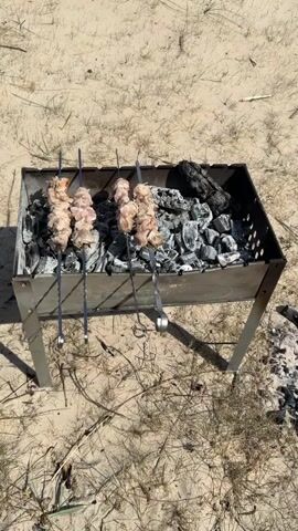 The video shows a barbecue grill on a sandy beach, where three skewers of meat are cooking over charcoal. In the background, a woman in a pink shirt is sitting in a green camping chair, recording a young girl who is blowing bubbles. The beach stretches to the sea.