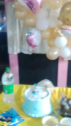 The video shows a party setup for a baby girl. There is a balloon arch with white and gold balloons. There is a baby girl balloon and two "Baby Girl" balloons as well.  Below the balloon arch there are two cakes on a yellow tablecloth. There is a bottle of Pepsi and some beer bottles as well. The background is a pink wall with a white curtain. There is also music playing in the background.