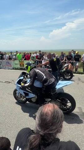 A medium shot shows two motorcycle racers and their bikes at a drag race. The racer on the left, with long hair and a black top, has their back to the camera, with their head in the lower-right corner. The first racer in the foreground wears a grey-and-black top, black pants, black shoes, and a yellow-and-black helmet, sitting on a light-blue-and-black sportbike. Another racer, on a grey-and-black sportbike, is further back on the right, being assisted by a person in a black shirt. A group of spectators stands behind a fence in the background. The race takes place on an asphalt road with green grass and trees on the horizon, under a partly cloudy sky.