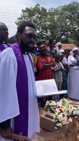 The video shows a funeral service taking place outdoors. A man dressed in clerical attire, holding a shovel, is reading from a document while standing next to an open grave. Behind him, a crowd of people, dressed in a variety of clothing, are gathered. The grave is lined with brick and stone, and a wooden coffin rests inside. A floral arrangement in the shape of a cross is placed on the edge of the grave. The atmosphere appears somber. The sky is overcast, and trees are visible in the background.