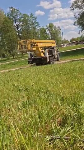 A yellow mobile elevated work platform on a six-wheeled truck is driving on a dirt road through a field of tall green grass.  The sky is blue with scattered white clouds and there are trees lining the background on either side of the road. A couple of houses are visible in the far distance.