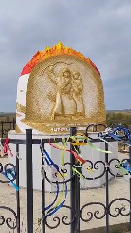 A golden relief of two figures in traditional clothing is set against a white, egg-shaped monument topped with fiery red and yellow. The figures are facing left; the one in front holds a raised arm. Below them, text is inscribed in gold. The monument is surrounded by a black metal fence, adorned with numerous colorful ribbons tied to its bars. The background shows a sparse, grassy landscape under a cloudy sky. The camera pans slightly to the right.