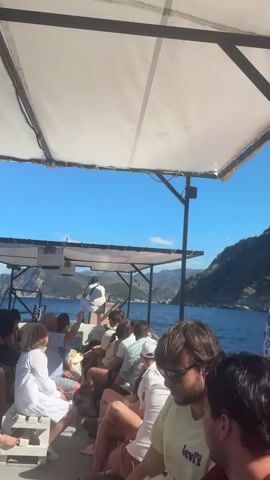 A video shows people sitting on a boat. The boat has a white canopy overhead, supported by black poles. In the background, there is a body of water and a lush green hillside under a clear blue sky. Several people are visible on the boat, some facing forward and others looking out at the scenery. One person stands at the front of the boat. The camera pans slightly, capturing the coastline and the passengers.