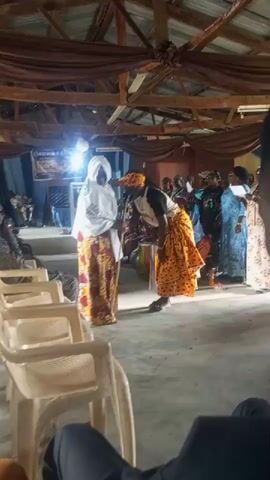 The video shows a gathering of people, possibly a church or community event, inside a building with a wooden ceiling and support beams. In the foreground, there are several rows of white plastic chairs. The main focus is on two women in the center of the frame. One woman, wearing a white headscarf and a colorful patterned skirt, stands facing a second woman. The second woman is also wearing a colorful patterned skirt and a dark shirt, with a head wrap. She is bent over, appearing to either tie the first woman's skirt or adjust it. Several other people are visible in the background, some standing and some seated. There is a stage area on the left with a banner and a sound system. The lighting is somewhat dim, with a bright spotlight on the stage area. The floor appears to be concrete.