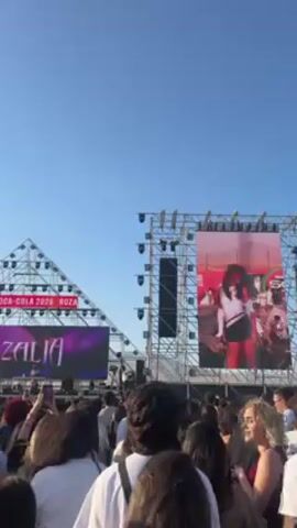 The video captures a concert or music festival scene from the perspective of someone in the crowd. The scene is outdoors with a clear, blue sky overhead. In the foreground, there is a large crowd of people, many holding up their phones to record. A stage is visible in the background, featuring a large screen displaying a performance by a woman with red hair, dressed in a white top and black skirt. The stage has a triangular structure to the left, displaying "COCA-COLA 2026" and "ROZALIA MUSIC FEST". The stage is also equipped with a significant amount of lighting equipment. The general atmosphere suggests a lively, crowded event.