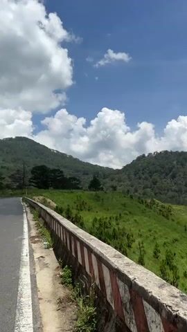 The video shows a scenic landscape of rolling green hills and lush forests under a bright blue sky with scattered white clouds. The camera pans from a roadside barrier, revealing the expansive landscape with dense forests covering the hills in the distance. There is a manicured golf course visible in the valley below. The overall environment is green and vibrant, suggesting a natural and possibly mountainous region.