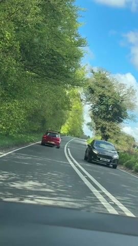 The video shows a first-person perspective from a vehicle driving on a paved road through a lush green landscape. A red hatchback car is visible ahead, driving in the same direction. The road has double white lines marking the center, indicating no overtaking. On both sides of the road, tall, verdant trees create a natural canopy and line the route. The sky above is bright blue with scattered white clouds, suggesting clear and sunny weather. To the right of the road, beyond the trees, a telephone pole is visible. The scene depicts a calm drive through a rural area in the Breckland District, United Kingdom, under clear daytime conditions.