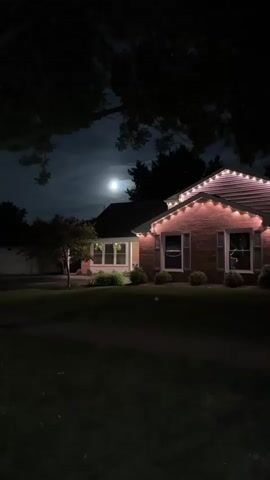 The video shows a night scene featuring a single-story brick house with warm-toned lights outlining its roof.  The house has multiple windows visible, and some are illuminated.  A neatly trimmed lawn covers the foreground.  A young tree stands to the left of the house. In the background, a full moon shines through a slightly cloudy sky, obscured somewhat by tree branches that frame the top of the scene. Another building is faintly visible to the left of the house. The scene is dark, but details are discernable, illuminated by the moon and the house lights.