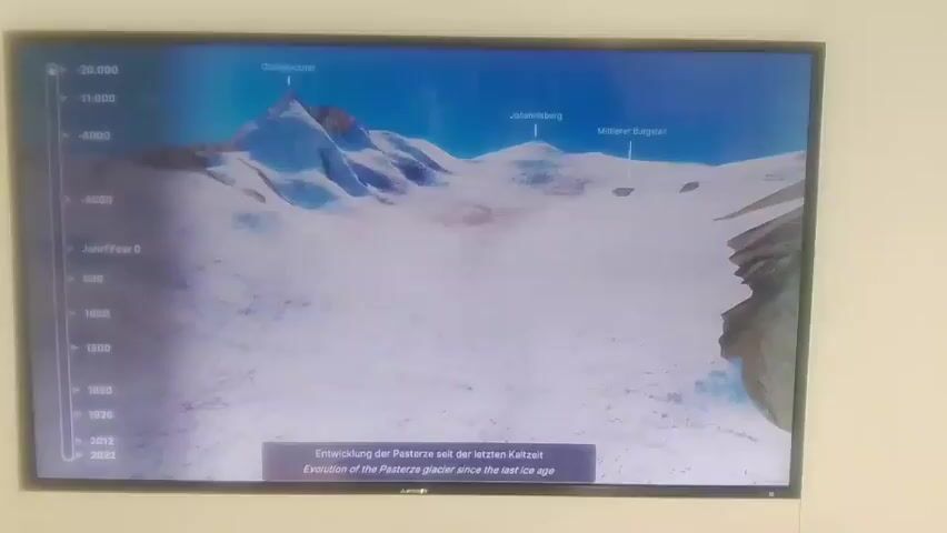 The video shows a television screen displaying a 3D animation of a glacier's evolution. A timeline is visible on the left side of the screen, with years ranging from the last ice age to 2022. The animation depicts the glacier receding over time, with prominent peaks labeled "Großglockner," "Johannesberg," and "Mittlerer Burgstall." The bottom of the screen displays text in German and English: "Entwicklung der Pasterze seit der letzten Eiszeit" and "Evolution of the Pasterze glacier since the last ice age." The screen is mounted on a white wall, and the lighting suggests an indoor environment. There are no people visible in the frame.