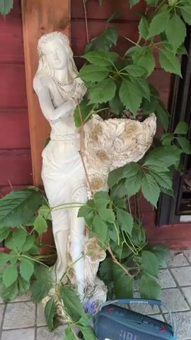 The video shows a white statue of a woman in a garden setting. The statue is adorned with jewelry and is partially covered by green vines. A large, ornate pot or planter sits next to the statue, also adorned with vines. In the foreground, a black and blue portable JBL speaker rests on the ground. The background consists of a reddish wooden wall and a tiled floor. The lighting suggests it is daytime, likely outdoors. The camera appears to be held by hand, as there is slight movement and refocusing throughout the clip.
