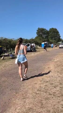 The video shows an outdoor market or car boot sale. A woman in denim shorts and a light-colored top walks away from the camera on a dirt path. Behind her, several cars are parked, and people are browsing items laid out on blankets or in car trunks. A blue portable toilet is visible in the background, along with trees and a clear blue sky. The ground is covered in dry grass and dirt. Another man is seen walking towards the right side of the frame carrying a yellow bag. A red SUV is parked in the foreground. In the distance, more cars and stalls are visible, suggesting a busy event.