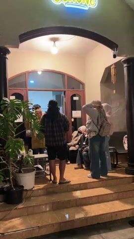 The video shows a group of people standing and sitting outside a building entrance at night. A neon sign above the entrance reads "GUNUNG" in green and yellow lettering. The entrance has double wooden doors and a large arched overhang. To the left of the entrance is a potted plant. The ground in front of the entrance consists of tiled steps. Several individuals are visible, some standing, others seated. One person in the foreground is wearing a plaid shirt and shorts. Another person in the foreground is wearing a light-colored jacket and jeans, with a backpack. The lighting is dim, suggesting it is evening or night.