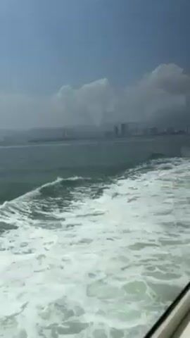The video shows a view from inside a moving boat. The camera is looking out through a window at the water and the wake of the boat. In the distance, there is a city skyline with tall buildings and mountains in the background. The sky is blue with white clouds.