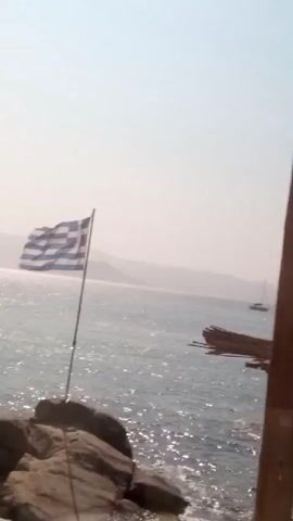 The video shows a slightly blurry shot of a coastal scene. In the foreground, a vertical wooden pole obstructs part of the view. To the left, a large Greek flag waves on a flagpole. The flag is positioned on a rocky outcrop near the water's edge. Beyond the flag, the sea stretches out, with sunlight glinting off the surface. In the distance, mountains are visible, silhouetted against a hazy sky. To the right of the frame, a rustic shelter with a thatched roof and wooden posts is partially visible, with some seating underneath. The water appears calm with small ripples. Further out at sea, a few small boats and a larger vessel can be seen. The overall environment suggests a sunny day at a seaside location, possibly a beach bar or a scenic viewpoint. The video appears to be filmed from a moving perspective, contributing to the slight blurriness.