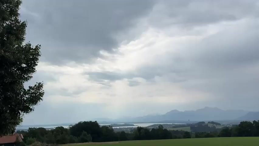 This is a wide shot of a cloudy, overcast sky over a hilly landscape with mountains in the distance. In the foreground, there is a green grassy field. To the left, there is a large tree with dark green leaves. In the middle ground, there are more trees and a body of water that appears to be a lake or a bay. Beyond the water, there are rolling hills and then the distant mountains, which are hazy and appear blue-gray. The clouds are dark and heavy, suggesting a potential for rain. There are no people or animals visible in the frame. The overall mood is somber and somewhat dramatic due to the weather.