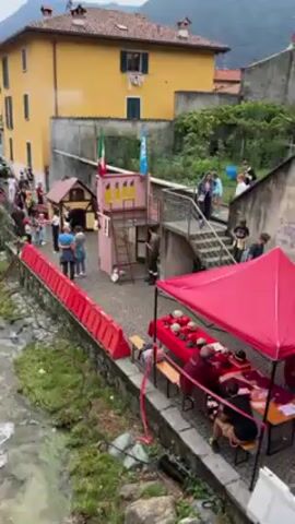The video shows a fast-flowing stream in a town setting with colorful buildings on either side. People are gathered along the side of the stream, near a small playhouse-like structure and a tent with tables. The scene includes an Italian flag, adding to the sense of place. A small building with a cross is located in the stream.