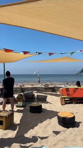 This is a medium shot video taken outdoors on a sunny day at a sandy beach. The video is framed by a tan shade sail stretched across the top and a string of colorful triangular flags. In the foreground, there is a sandy area with various beach furniture, including lounge chairs, circular tables made from tires, and wooden crates. A person in a black shirt and shorts stands on the left, facing away from the camera. In the middle ground, a wooden barrier separates the seating area from the beach. Beyond the barrier, people are sunbathing and walking on the sand. The ocean stretches across the background with gentle waves and a clear blue sky above. The main action of the video is a person standing on a tall pole near the water's edge, holding a flag. The person appears to be adjusting or interacting with the flag, which is yellow and white with a graphic design on it. The person is wearing red shorts and is barefoot. Several other people are visible on the beach and in the water, some near jet skis. The overall atmosphere is relaxed and summery.