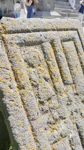 The video shows a close-up of an ancient stone carving covered in lichen. The carving appears to be a rectangular pattern with raised lines and some sort of symbol or inscription in the center. In the background, out of focus, there are people walking and standing near some stone steps, suggesting this is an outdoor historical site. The lighting indicates it is daytime.