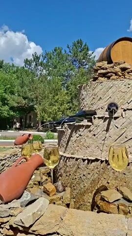 A fountain made of stacked stone with wine bottles and glasses is displayed in a park. Water flows from the bottles and into the glasses. A wooden barrel sits on top of the fountain. Trees and a blue sky with clouds are visible in the background.