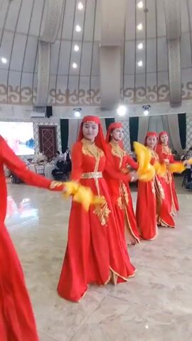 Four women in traditional red and gold dresses with head coverings are performing a dance in a large hall with a domed ceiling. They are holding yellow fans and moving in unison. The hall has a polished floor and a screen in the background showing more dancers. There are tables and chairs set up in the background.