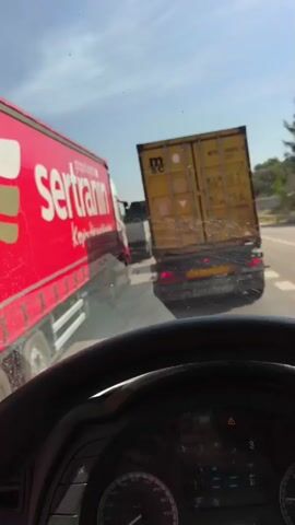 The video shows a view from inside the cab of a truck, looking out at heavy traffic. The truck is surrounded by other large trucks, including one with the "sertranin" logo on a red trailer, and another with an "MSC" container on a yellow trailer. The side mirror of the truck reflects the red "sertranin" truck and the vehicles behind it. The dashboard and steering wheel of the truck are visible. The sky is blue and sunny.