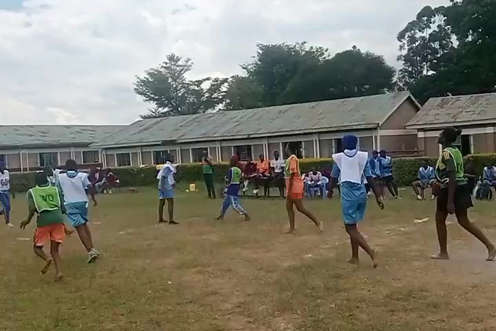 The video shows a group of young people playing a game on a grassy field, possibly netball. Most of them are wearing light blue shorts and white tops, some with colored vests over them. They are moving around the field, passing and attempting to catch a red ball. In the background, there is a long, low building with a green roof, and several people are sitting and watching the game. The sky is partly cloudy.
