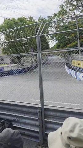 A racing car drives down a paved track. Spectators are watching the race from behind a fence. The sky is cloudy.