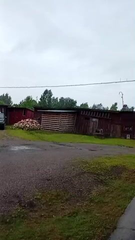 The video shows an outdoor scene on a cloudy day, possibly raining, as indicated by reflections on the ground and some droplets visible in the air. In the foreground, there is a gravel path with some wet patches and grass bordering it. To the left, there is a pile of what appears to be lumber or wood. Behind this, a long, weathered, red-colored building is visible, featuring a wooden structure with horizontal slats. Further to the right, the building continues, showing signs of age with some exposed wood and metal siding. There are windows and doors on the building. A power line stretches across the scene above the building. In the distance, trees form a treeline behind the structures. A satellite dish is mounted on a pole near the right edge of the frame, extending upwards. The top right corner of the video shows a portion of an overhang or roof structure, possibly part of the viewpoint.
