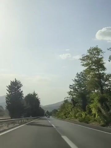 The video shows a point-of-view shot from a moving vehicle on a highway. The road is paved with a white dividing line. On the left side of the road, there is a metal guardrail and a line of trees, some of which are tall and coniferous. On the right side, there are more trees and vegetation. In the distance, mountains are visible under a hazy sky with scattered clouds. A few other vehicles are seen on the road ahead. The lighting suggests it is daytime, possibly late morning or afternoon, with the sun high in the sky, creating some glare.