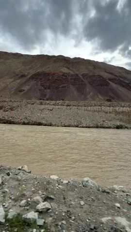 This is a video recording of a wide, muddy river flowing through a mountainous landscape under a cloudy sky. The camera is positioned low, near the riverbank, which is composed of rocks and dirt. The mountains on the opposite bank are steep and barren, with visible rock formations and varying shades of brown and purple. Sparse vegetation, including small trees and bushes, can be seen along the riverbanks and on the lower slopes of the mountains. The river itself appears to be flowing at a moderate pace, with ripples and some debris visible on its surface. The sky is overcast with dark, dramatic clouds, suggesting the possibility of rain. The video pans slightly to the right, revealing more of the river and the surrounding mountainous terrain. There are no people or vehicles visible in the video.
