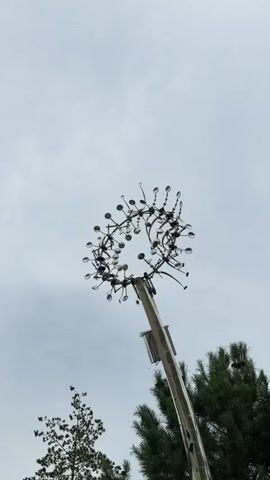 The video shows a low-angle shot of a kinetic sculpture mounted on a pole against a cloudy sky. The sculpture is circular, with numerous thin, flexible rods extending outwards from a central ring. Each rod is tipped with a small, reflective sphere. Some rods are bent and curved, giving the sculpture a dynamic, almost organic appearance. The pole supporting the sculpture is visible below it, and two trees, one with sparse foliage and the other a conifer, are partially visible in the background. The sculpture appears to be moving gently, as evidenced by the subtle shifts in the positions of the rods and spheres. The overall impression is one of outdoor art or a wind-activated installation.