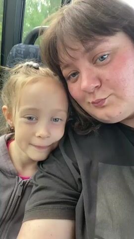 This is a close-up, selfie-style video featuring two individuals, likely a mother and daughter, seated inside a vehicle. The woman on the right has medium-brown hair and fair skin with a scattering of freckles. She is wearing a dark gray t-shirt and is looking directly at the camera with a slight pout. The girl on the left has blonde hair pulled back and is wearing a gray zip-up hoodie over a pink top. She is also looking at the camera, with a slightly pouting expression, her head leaning against the woman's shoulder.

The setting appears to be the interior of a bus or van, suggested by the pattern on the headrest visible behind the subjects. Through the window to the left, a blurred green, natural landscape is visible, hinting at an outdoor setting or a journey through a rural area. The lighting suggests it is daytime. The overall mood of the video is warm and affectionate, with the close proximity and shared expressions indicating a bond between the two individuals. There is no visible text in the media.