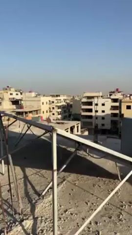 The video shows a view from a rooftop in what appears to be a war-torn urban environment. In the foreground is a metal frame structure, possibly for solar panels, on the rooftop. In the background, there are several multi-story buildings, some of which are heavily damaged. One building in particular has significant structural damage, with large sections missing and debris visible. The sky is clear and the lighting suggests it is daytime.  No people are visible in the video.