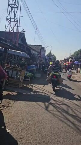 The video shows a street scene with several people on motorcycles and scooters. The street is lined with market stalls on both sides, displaying various goods. Sunlight filters through the scene, casting long shadows on the asphalt road. A few people are visible walking among the stalls. The overall atmosphere appears to be that of a bustling local market on a sunny day.