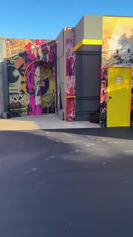 A digital video displays an outdoor setting with a backdrop constructed of various panels, some featuring large-scale graphic designs. The graphic elements include a stylized portrait of a woman, bold abstract shapes in pink, yellow, orange, and black, and elements resembling torn paper collage. On the left side of the backdrop, there is a gray panel with a curved metal section. To the right, the backdrop is predominantly gray with a bright yellow door. In the foreground, a vibrant yellow sofa with two pink cushions is placed on a dark gray surface. Two small, dark-colored side tables with round tops are positioned on either side of the sofa. In the lower portion of the frame, camera equipment, including cables and a spotlight, is visible on a gray concrete surface. The scene is illuminated by natural sunlight, casting shadows across the ground. The sky is a clear blue.
