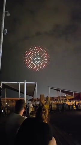 The video shows a drone light show at night. A large ring of red and white lights is visible in the sky, forming a circular pattern. Below the drone display, there are people seated, facing away from the camera. Structures with awnings and lights are visible in the background, suggesting an outdoor event venue. The sky is dark with some clouds. The overall scene captures a moment of spectacle and entertainment.