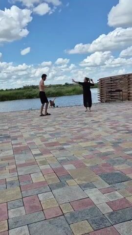 Two people are dancing on a paved area near a body of water. A man in black shorts and no shirt is in a dynamic pose, possibly mid-dance move. A woman in a black dress is also dancing, facing the man. Behind them, a wooden structure with a roof is visible. The sky is blue with scattered white clouds. The camera pans to show other people near the water, one man in a patterned shirt and shorts is standing, while another person is hunched over. The camera then moves to a swimming pool area, where a person is in the water. Another wooden structure is present near the pool, and several pairs of flip-flops are scattered on the ground. The scene appears to be outdoors on a sunny day.