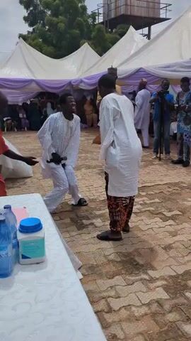 In an outdoor event, two young men are in the center of the frame. The man on the left is wearing a white traditional outfit with intricate embroidery. He is in a dynamic pose, leaning forward with his arms extended and his head tilted back as if shouting or laughing. The man on the right is wearing a white traditional top and patterned pants. He is facing the first man and holding a microphone close to his face, seemingly interacting with him. A third person in a red outfit is partially visible on the far left, also appearing to be interacting with the man in white. In the background, there are white tents with purple drapes, suggesting a festive or celebratory occasion. Other people are visible further back, some holding cameras. A table in the foreground on the left holds several bottles of water and a container with a blue lid. The ground is paved with interlocking bricks. The sky is cloudy.