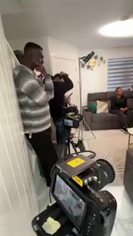 The video shows a scene from what appears to be a film shoot in a living room setting. A woman sits on a gray couch, seemingly the subject of the scene. Several people are present, including crew members operating cameras and adjusting equipment. One person, wearing an orange shirt, stands near the woman, possibly giving direction or interacting with her. Another woman sits on a different couch, observing the scene. The room is decorated with a patterned rug, side tables, and modern furniture. The overall impression is a behind-the-scenes glimpse of a production in progress.