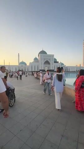 A person in a wheelchair is being pushed by another person across a paved area in front of a large white mosque with turquoise domes and minarets. Several other people are walking or sitting in the background, some standing in a line near a body of water. The sky is clear and blue, suggesting it is daytime. One woman in the foreground is taking a picture with her phone.