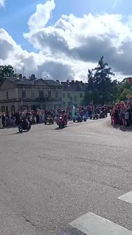 A group of people are watching a parade on a street lined with buildings. The parade consists of motorcycles and ATVs, some of which are emitting smoke. One ATV in the center of the frame is carrying a flag. The sky is blue with some clouds.