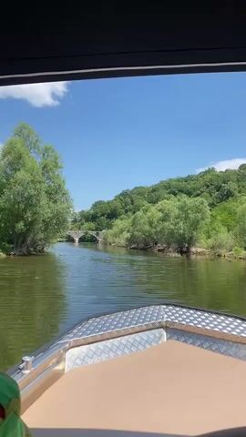 The video shows a first-person perspective from a boat moving down a river on a sunny day. The boat is a small, modern vessel with a tan seat and a metallic bow. Lush green trees line both banks of the river. In the distance, an old stone bridge with multiple arches is visible, spanning the river. The sky is blue with a few white clouds. The water appears calm and reflects the surrounding greenery.