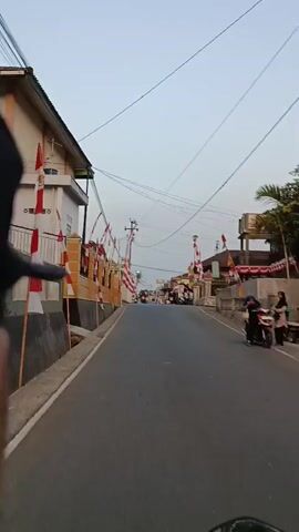 The video is shot from a first-person perspective, likely from a motorcycle, moving down a street lined with buildings decorated with Indonesian flags. The flags, red and white, are strung along both sides of the road, creating a festive atmosphere. There are several motorcycles visible on the road, both in front and in the distance. The sky is clear and appears to be dusk. In the foreground, the top of a black cap is visible, suggesting the person recording is wearing one. The buildings on the left are light yellow with white trim, and a gray wall runs along the sidewalk. The road surface is asphalt. Electrical wires are strung overhead. The overall impression is a quiet street scene, possibly in preparation for or during a national celebration.