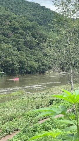 A group of people are in a pink raft on a river, surrounded by lush green trees and a mountain. The water appears calm and the sky is partly cloudy. The raft is moving downstream and the people seem to be enjoying a leisurely ride. The vegetation on the riverbank is dense and green.