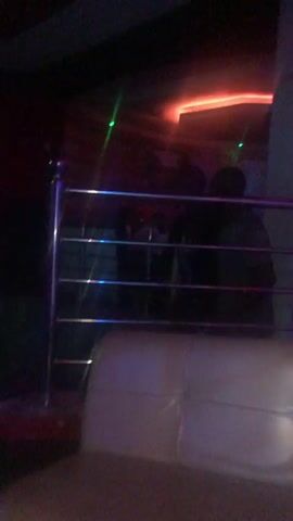 The video shows a blurry, dimly lit interior scene, likely a club or bar, with changing colored lights, primarily blue, red, and green. A metal railing is visible in the foreground. In the background, indistinct figures can be seen. The camera appears to be moving, giving a sense of being in motion within the space. There are no clear actions or interactions captured due to the blurriness and lighting.