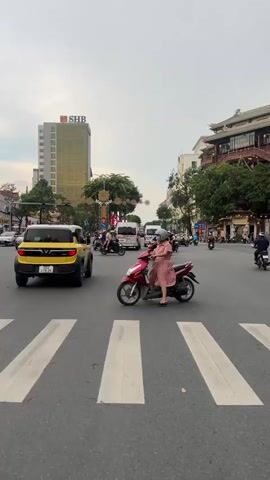 A street scene in Vietnam. A yellow car is driving down the street, and a woman is riding a motorcycle. There are many other motorcycles and cars on the street as well. The buildings on either side of the street are tall and modern, with some traditional architecture mixed in. The sky is cloudy.