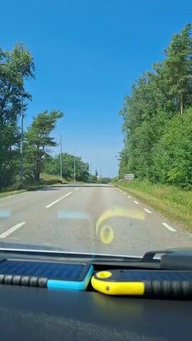 The video is shot from the perspective of a driver looking through the windshield of a car. The car is driving on a paved road with a clear blue sky above. There are trees lining both sides of the road, and some streetlights are visible. A white car is seen driving in the opposite direction. In the distance, a church steeple is visible above the trees. A sign is also visible on the right side of the road. There are two electronic devices on the dashboard in the foreground, one blue and one yellow.