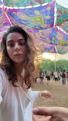 A woman with curly hair, wearing a white top, is dancing at what appears to be a music festival. Behind her, other people are also dancing and walking around. Above the crowd is a large, colorful canopy featuring psychedelic patterns. In the foreground, a person gives a thumbs up while recording the video. The ground is sandy and the general atmosphere seems relaxed and festive.