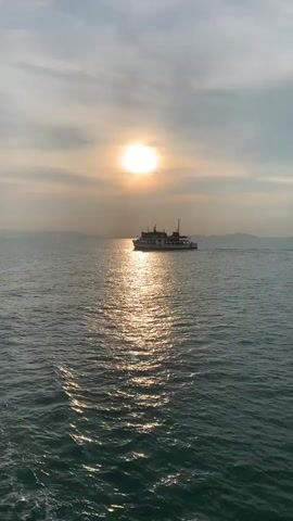 The video shows a ferry traveling across a body of water, silhouetted by the sun. The sun is high in the sky, with its light reflecting off the water, creating a shimmering effect. In the background, there are hints of land. The ferry, labeled "SEATRAN 6," is moving from left to right in the frame.